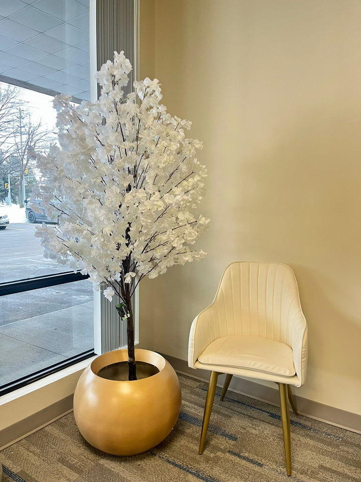 Artificial cherry blossom tree featuring soft white petals and branching structure, placed beside a fabric chair on light wooden flooring.