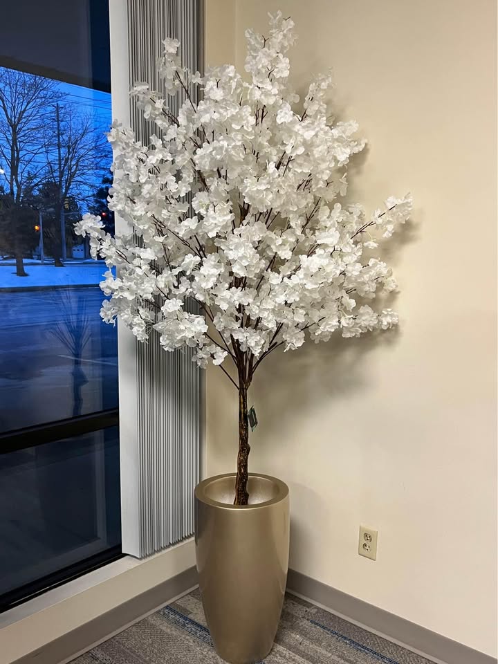Tall artificial white cherry blossom tree positioned near a large window in a contemporary apartment corner setting.