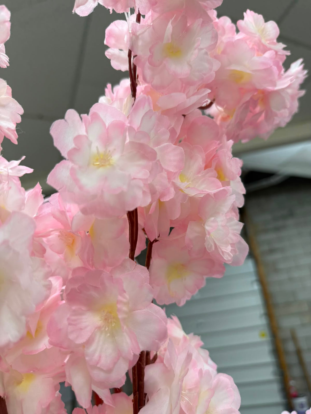 Pink artificial cherry blossom tree branch with multiple clusters of soft pink flowers and yellow centers on a brown stem, photographed close-up against an indoor background.