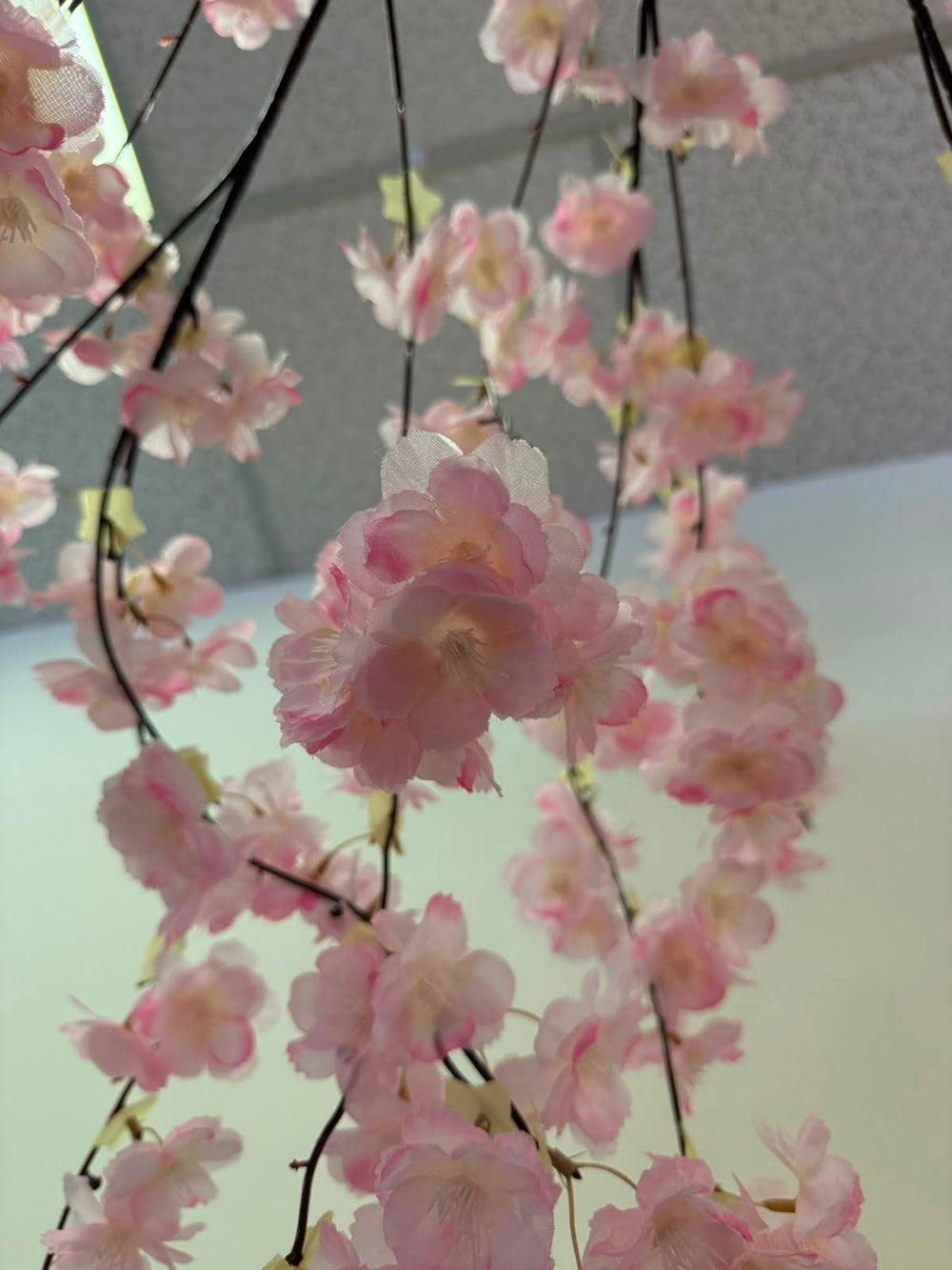 Pink artificial cherry blossom branches hanging down, featuring soft pink flowers with dark brown stems against a light gray and white background.