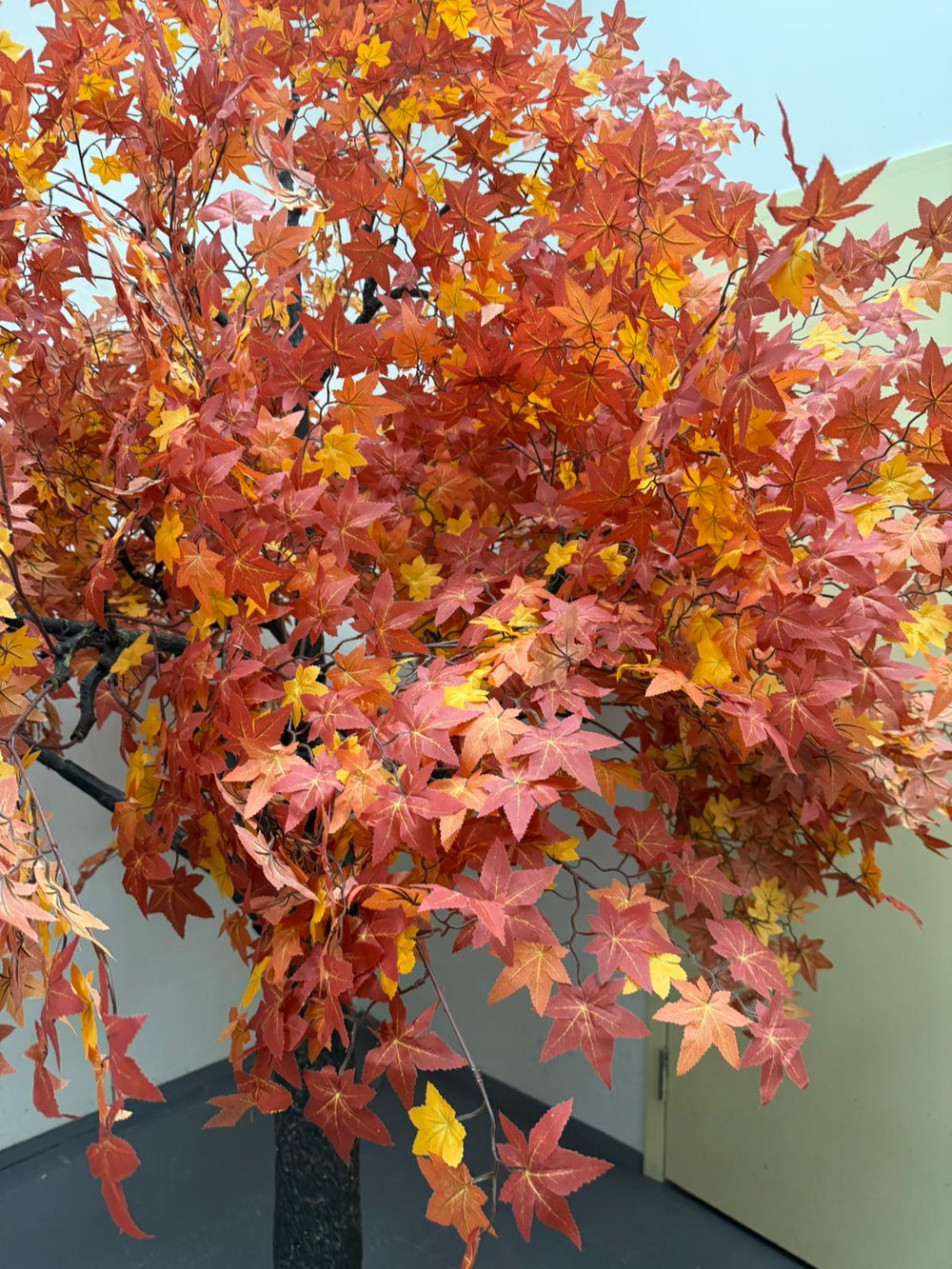 An artificial autumn maple leaf tree with dense branches covered in red, orange, and yellow leaves, shown indoors against a plain wall.