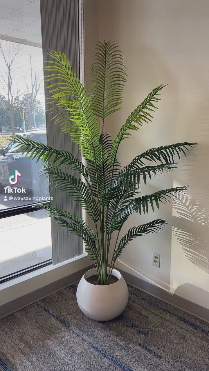 A stylish artificial palm tree placed in a white, round planter, adding a touch of greenery and elegance to this office or living space near a window. The natural light accentuates the realistic appearance of the lush, green fronds, creating a serene and inviting atmosphere.