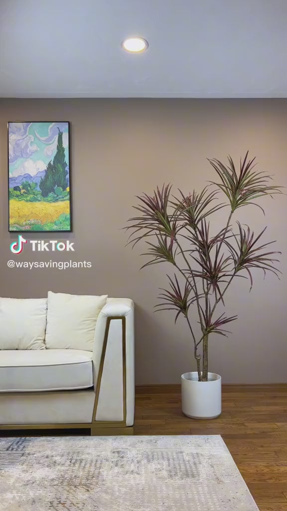 Contemporary living room interior featuring a white sofa with gold accents and a faux Dracaena plant with red-tinted leaves in a white planter. The artificial plant complements the minimalist decor, alongside a vibrant wall painting, adding a touch of color and elegance to the space.