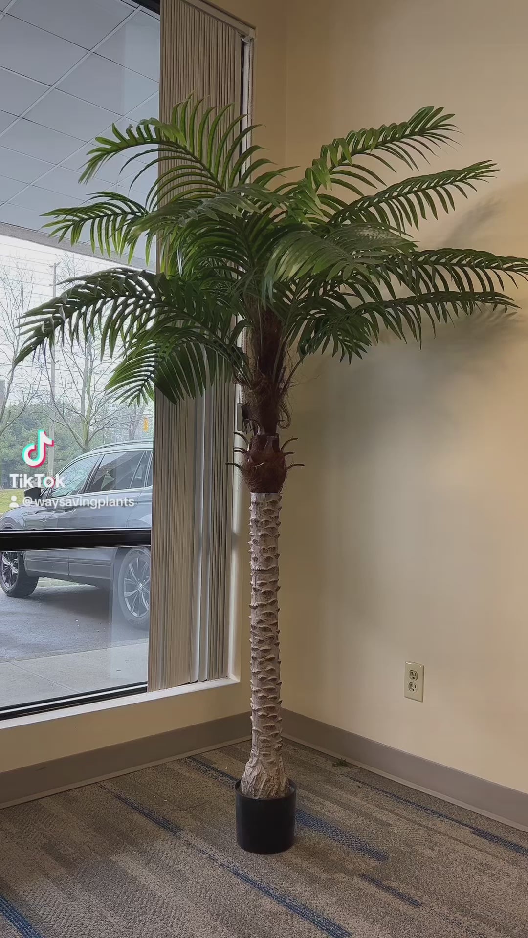 A large artificial palm tree in a stylish office corner by the window, adding a natural touch to a professional setting.