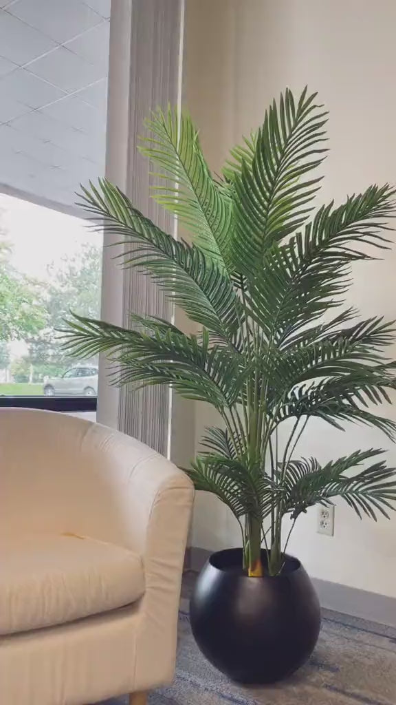 Artificial palm tree with green fronds, placed in a sleek black pot next to a white sofa, adding a natural touch to a modern, well-lit living room.