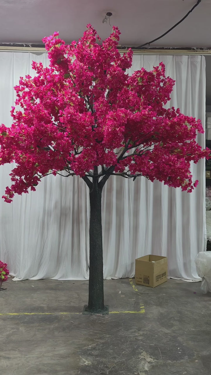 A vibrant artificial bougainvillea tree with lush pink flowers on a dark green trunk, displayed indoors against a white draped backdrop. The scene includes cardboard boxes and work materials in the background, suggesting a workshop or preparation area.