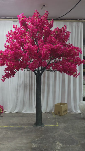 A vibrant artificial bougainvillea tree with lush pink flowers on a dark green trunk, displayed indoors against a white draped backdrop. The scene includes cardboard boxes and work materials in the background, suggesting a workshop or preparation area.