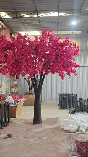 Elegant artificial tree adorned with red blossoms, featured in a beautifully set event hall with round tables, floral centerpieces, and wooden chairs. Can these decorative trees be customized to match specific event themes?