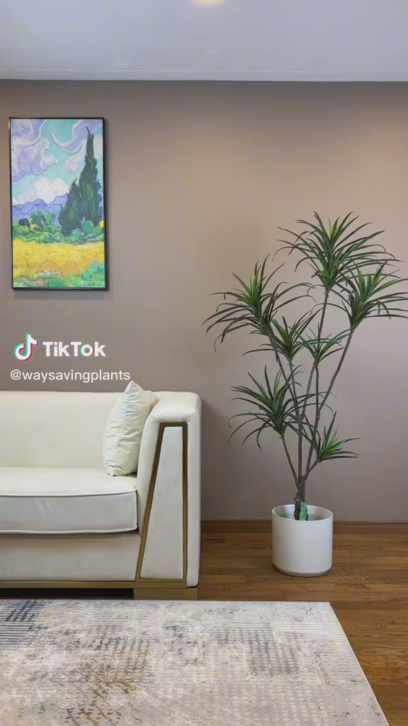 Artificial yucca plant in a modern white planter placed beside a luxurious white sofa with gold accents, enhancing the contemporary decor of a living room with wooden flooring and wall art