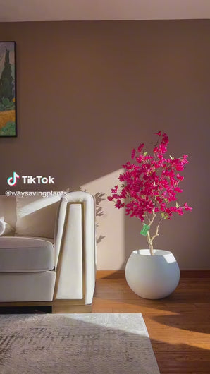 Artificial pink flowering tree in a white pot, placed beside a cream-colored couch with sunlight streaming in, highlighting the vibrant colors in a cozy living room setting