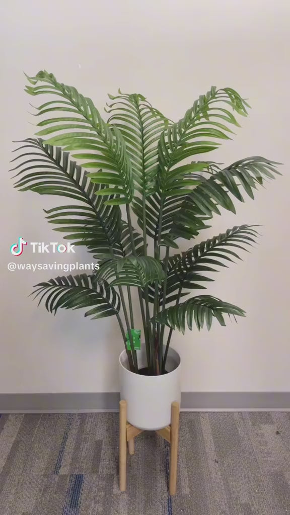 Artificial palm tree in a modern white pot with a wooden stand, displayed against a neutral wall and carpeted floor, ideal for indoor décor.