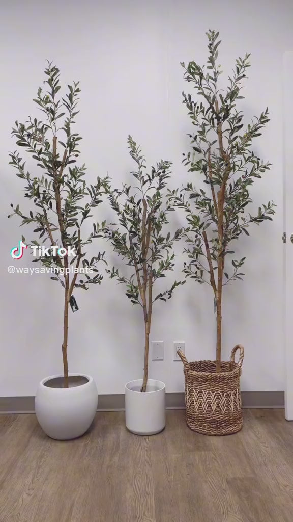 Three artificial olive trees of varying heights are displayed indoors, each in a different container—one in a round white pot, one in a cylindrical white pot, and the third in a woven basket.