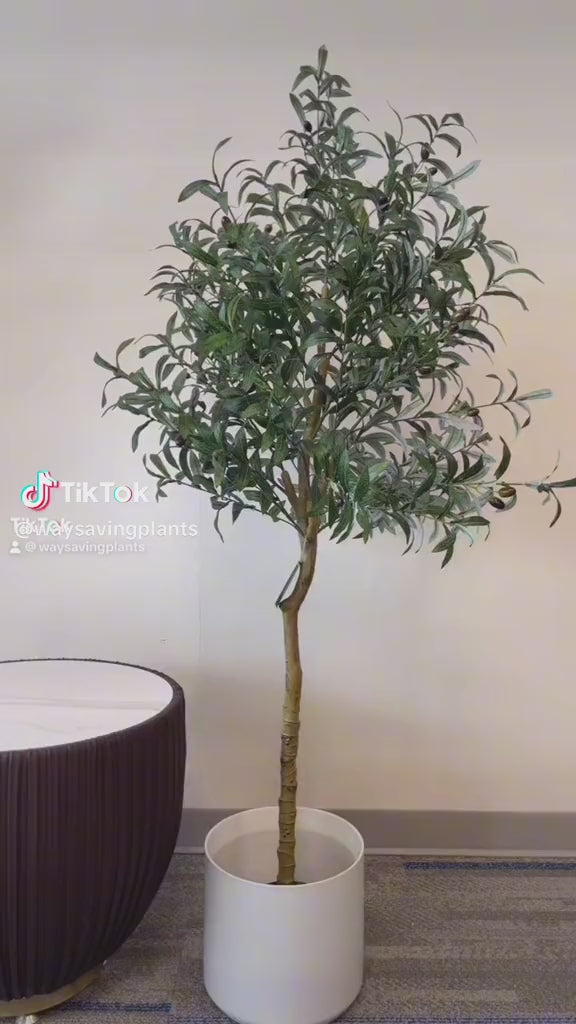 Artificial olive tree with realistic green leaves and black olives in a sleek white pot, positioned next to a modern, dark-colored round side table with a white top, placed on a light-colored carpet in an indoor setting