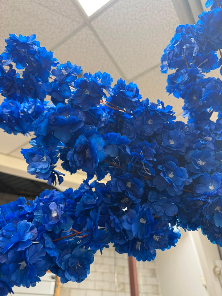 A close-up of a decorative artificial flower  branch with dense clusters of vivid royal blue blossoms, photographed indoors against a ceiling tile and wall background.