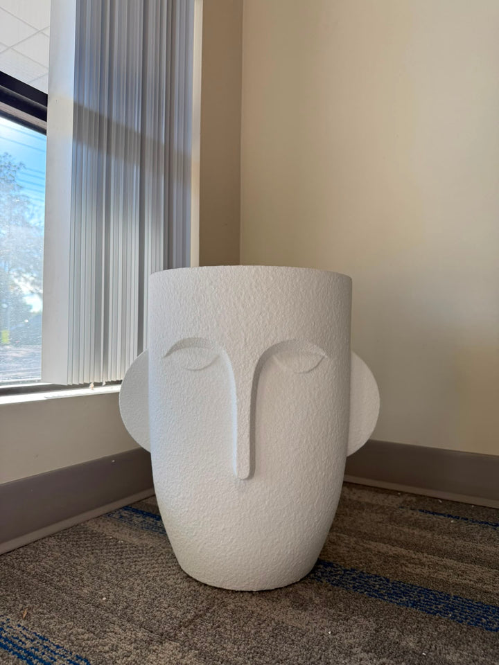 A modern white textured planter shaped like a stylized face, placed on a patterned carpet in a room corner next to a window with blinds.