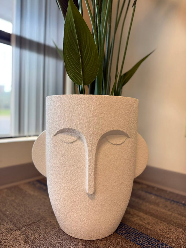A sculptural off-white planter shaped like a stylized face with closed eyes, displayed on a carpeted floor near a window with a green plant inserted.