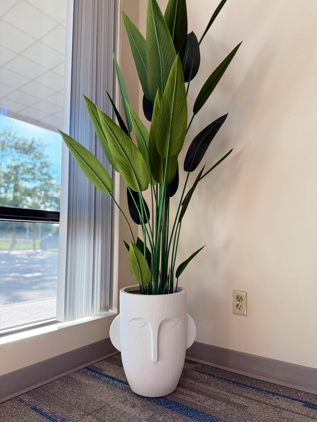 A tall artificial plant with green and dark green leaves in a white sculptural  pot, placed in the store next to a window.