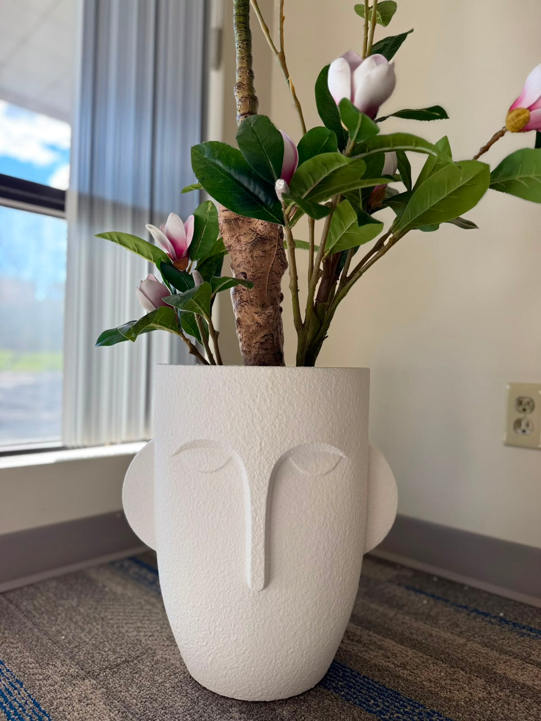 A white textured planter shaped like a stylized face, filled with green leaves and pink-and-white magnolia flowers, placed on a carpeted floor near a window.
