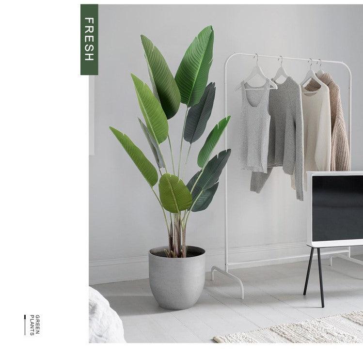 A stylish interior featuring an artificial bird of paradise plant in a sleek gray planter. The plant's large green leaves add a fresh, vibrant touch to the minimalist space. Nearby, a white clothing rack displays neutral-toned sweaters and tops, contributing to the room's clean, modern aesthetic. The muted color palette and simple furnishings create a serene and contemporary environment