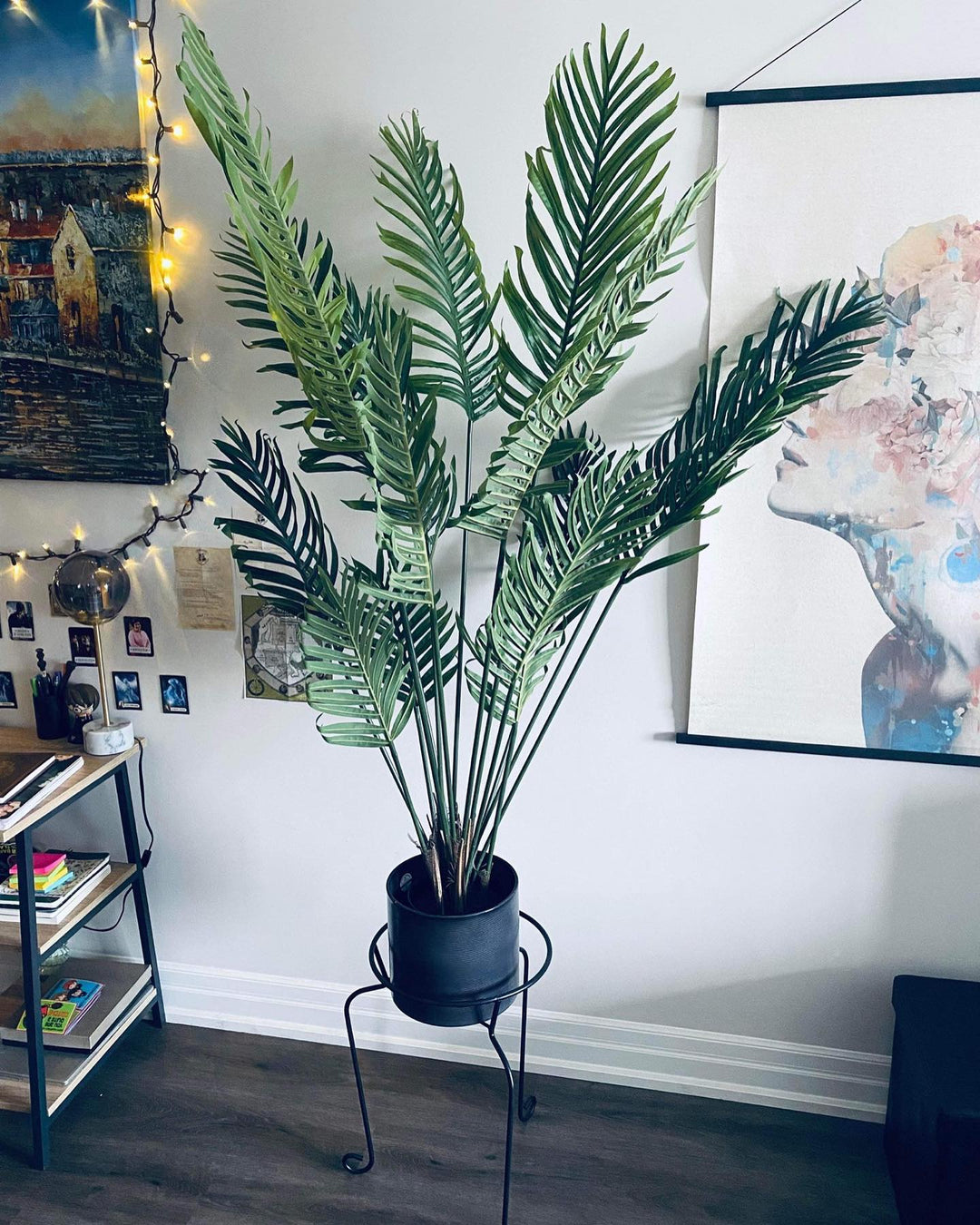 An artificial palm tree placed in a black pot, elevated on a metal plant stand. The tree's vibrant green leaves fan out gracefully, complementing the surrounding decor, which includes a framed wall art piece and string lights. The setup adds a lively and cozy touch to the room.