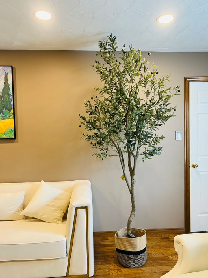 big fake olive trees. A cozy living room featuring a tall artificial olive tree in a stylish woven basket planter, placed next to a cream-colored modern sofa against a warm beige wall