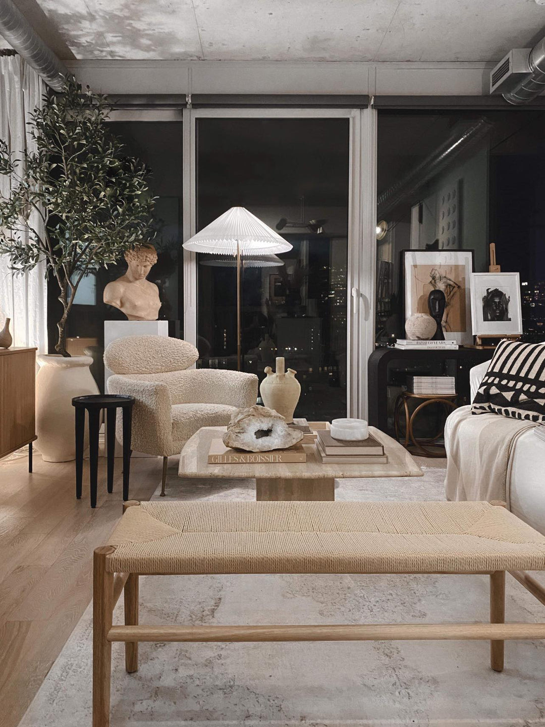 A stylish, modern living room with floor-to-ceiling windows at night, featuring a tall artificial olive tree, cozy textured armchair, and a mix of contemporary and classic decor, including a bust statue, wooden bench, and a soft neutral color palette