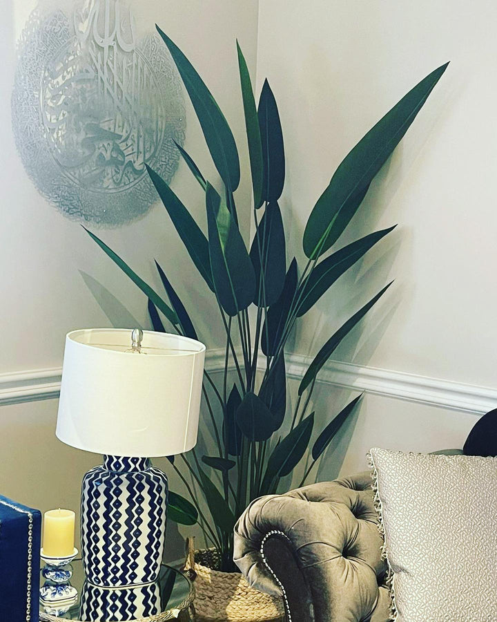 A stylish living room corner featuring a tall artificial bird of paradise plant with long, slender leaves placed beside a tufted beige armchair. A decorative blue and white patterned lamp sits on a glass-topped side table, adding a modern touch to the space. The room is accented with wall art and subtle, neutral decor, creating an inviting atmosphere.