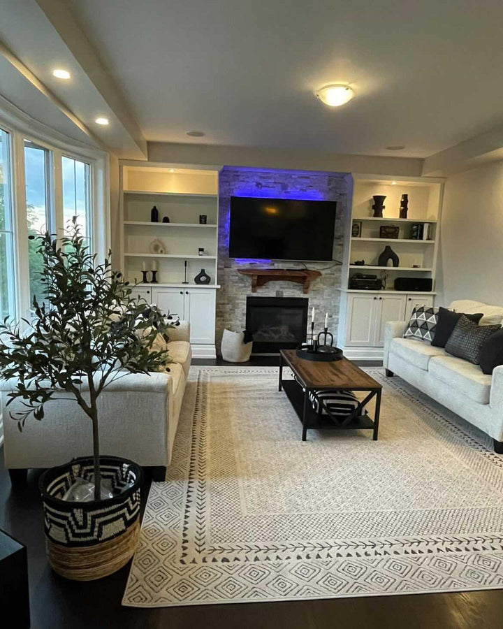 Modern living room interior featuring a large, cozy white sectional sofa and a stylish artificial olive tree in a black and white patterned basket. The room is designed with a neutral color palette, a light beige area rug with geometric patterns, and built-in shelving around a stone fireplace with a mounted TV above it. The space is illuminated by recessed ceiling lights and large windows that allow natural light to fill the room. The olive tree adds a touch of greenery, enhancing the contemporary.