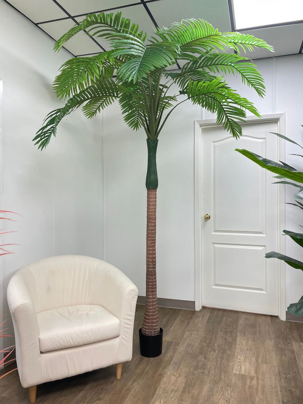Contemporary office space with a tall artificial palm tree beside a plush white armchair, creating a relaxing corner in a clean, minimalist setting.