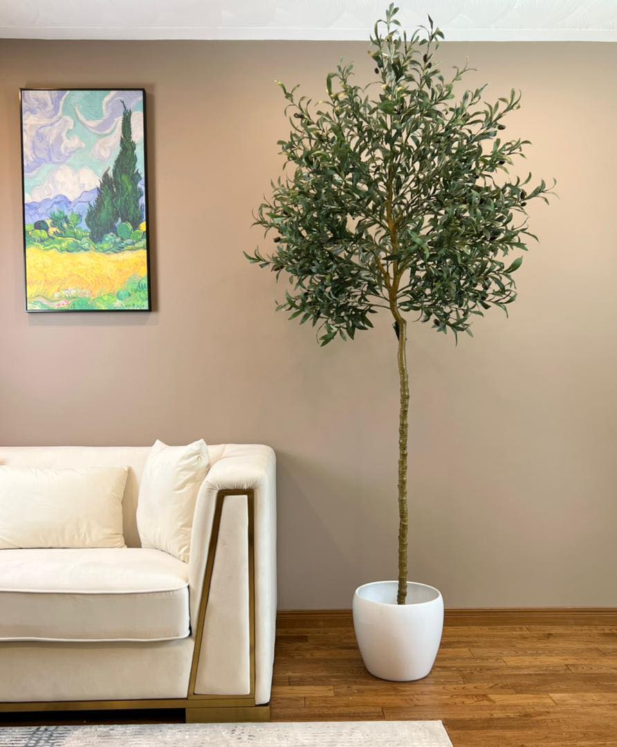 Elegant living room setup featuring a tall artificial olive tree in a white pot positioned next to a modern beige sofa with gold trim. The room has a warm, brown-toned wall adorned with a colorful landscape painting, creating a cozy and sophisticated atmosphere