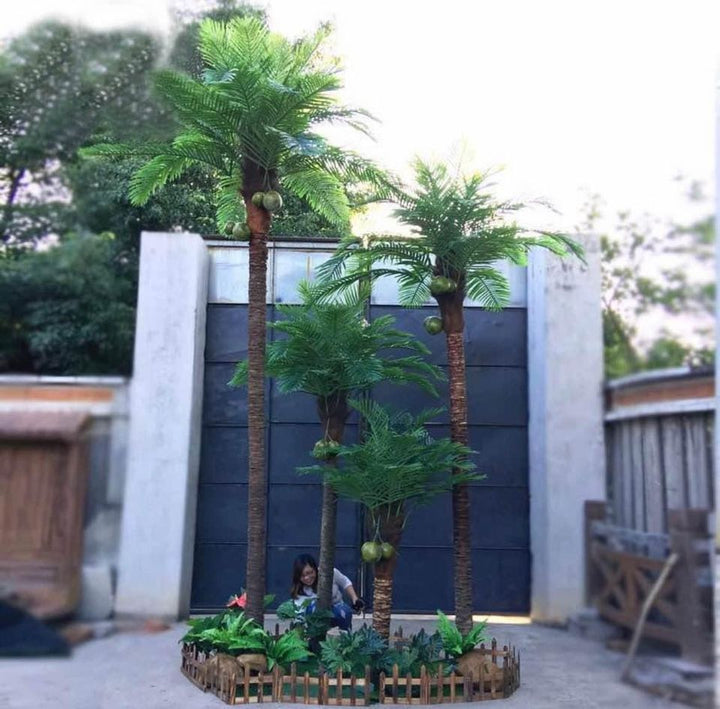 The image shows a cluster of artificial palm trees with coconuts in a small, fenced garden bed. The setup is outdoors, against a large dark gate, creating a tropical vibe.