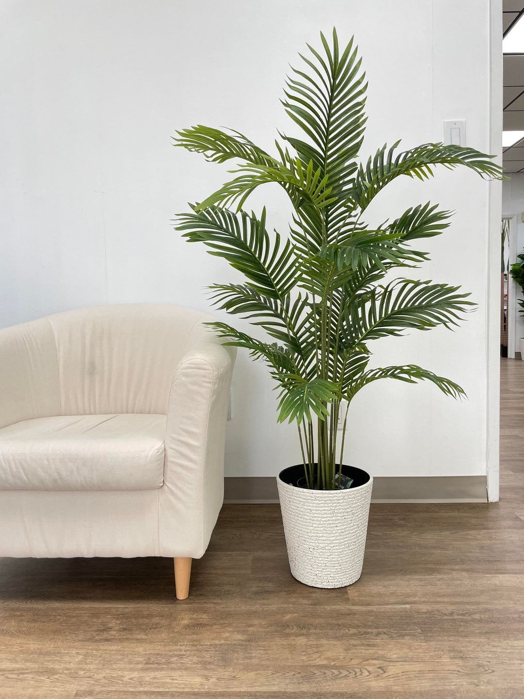 Minimalist office decor featuring a lush green palm plant in a white woven pot, placed next to a comfortable beige armchair on a wooden floor.