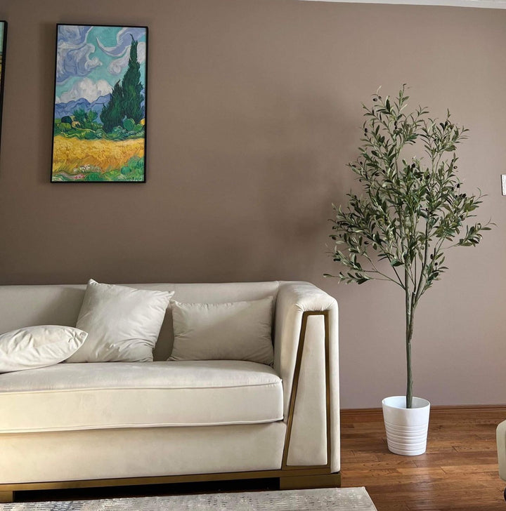 Elegant living room setup featuring a tall artificial olive tree in a white pot positioned next to a modern beige sofa with gold trim. The room has a warm, brown-toned wall adorned with a colorful landscape painting, creating a cozy and sophisticated atmosphere