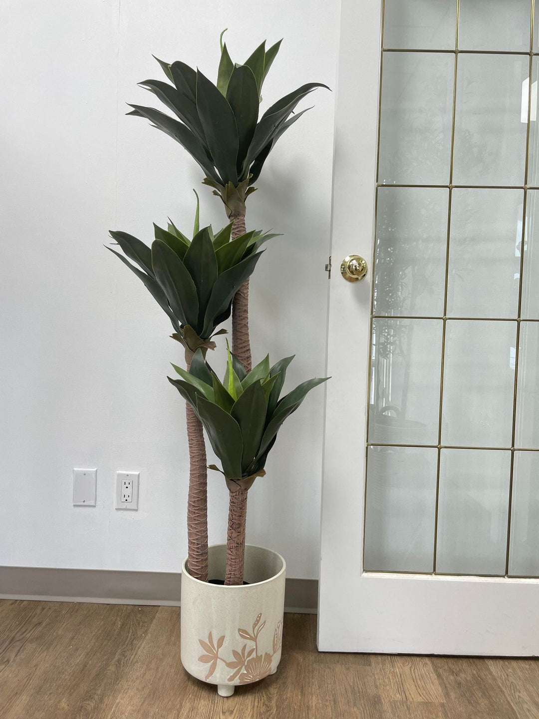 Modern living room setup featuring a cream-colored sofa with a faux yucca plant in a white pot next to a door. The artificial plant adds greenery to the minimalist decor, enhancing the stylish interior design.