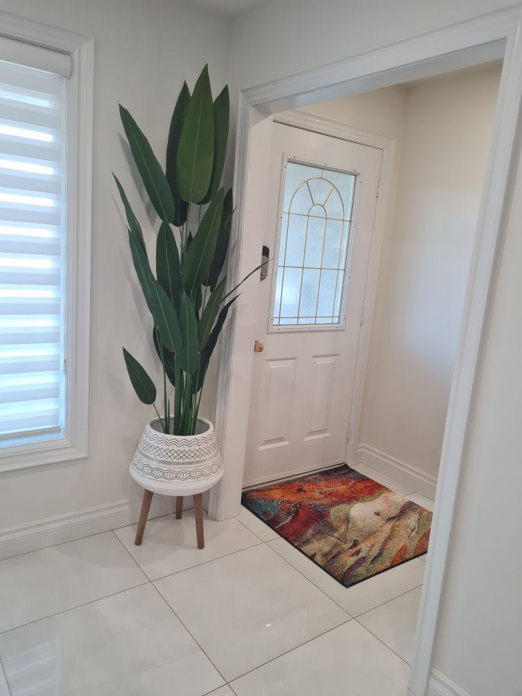 A bright and welcoming entryway featuring a tall artificial bird of paradise plant in a decorative white planter with wooden legs. The plant is placed next to a door with a frosted glass window, adding a touch of greenery to the clean, white-tiled space. A colorful rug with abstract patterns enhances the warmth and vibrancy of the entrance area