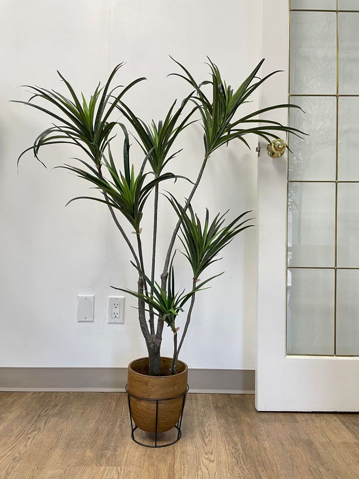 Artificial Dracaena tree in a brown planter with black metal stand, placed against a white wall and wooden floor, perfect for indoor home or office decor