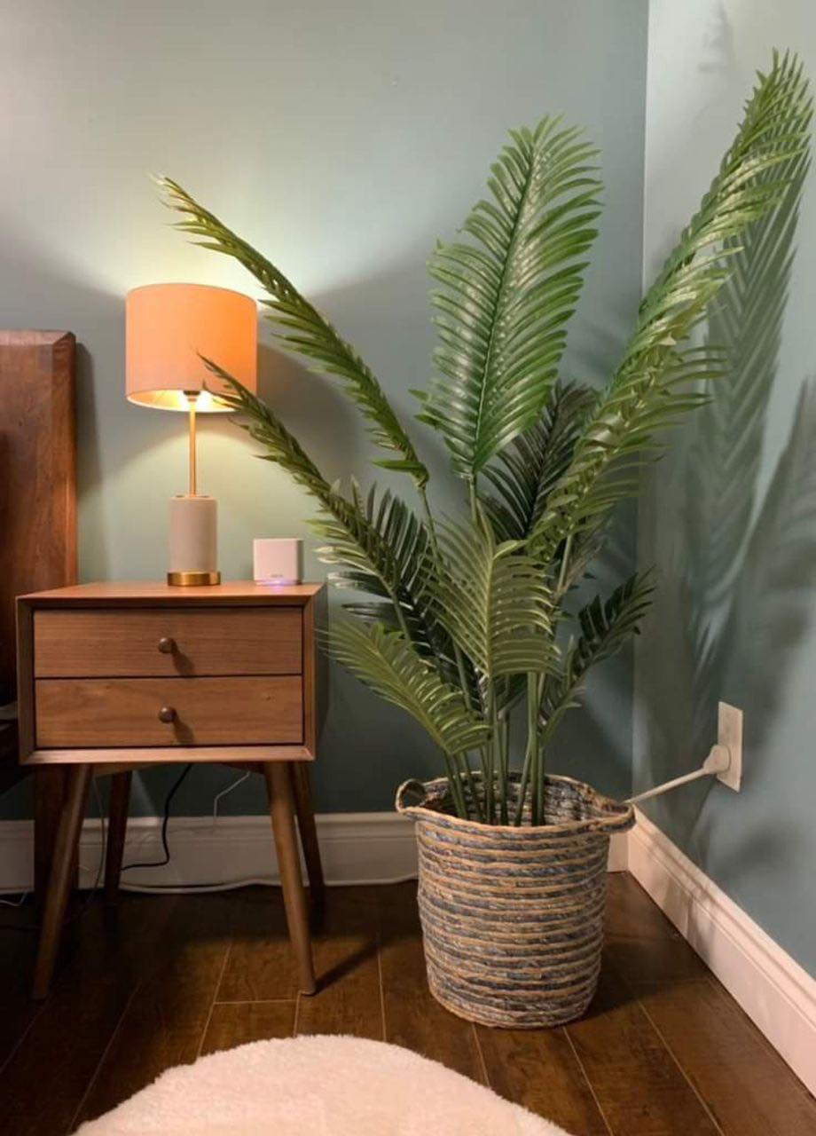 A cozy bedroom corner featuring an artificial palm tree in a woven basket. The setup includes a wooden side table with a warm-toned lamp, adding a soft glow to the room. The light green walls and the natural textures create a calming and serene ambiance.