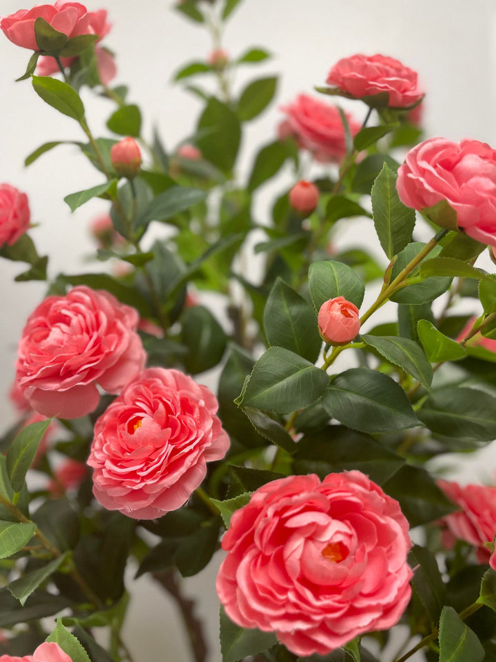 This image showcases a close-up of vibrant pink artificial flowers, surrounded by lush green leaves. The petals are layered delicately, mimicking the natural beauty of real flowers. The background is softly blurred, emphasizing the flowers' texture and color