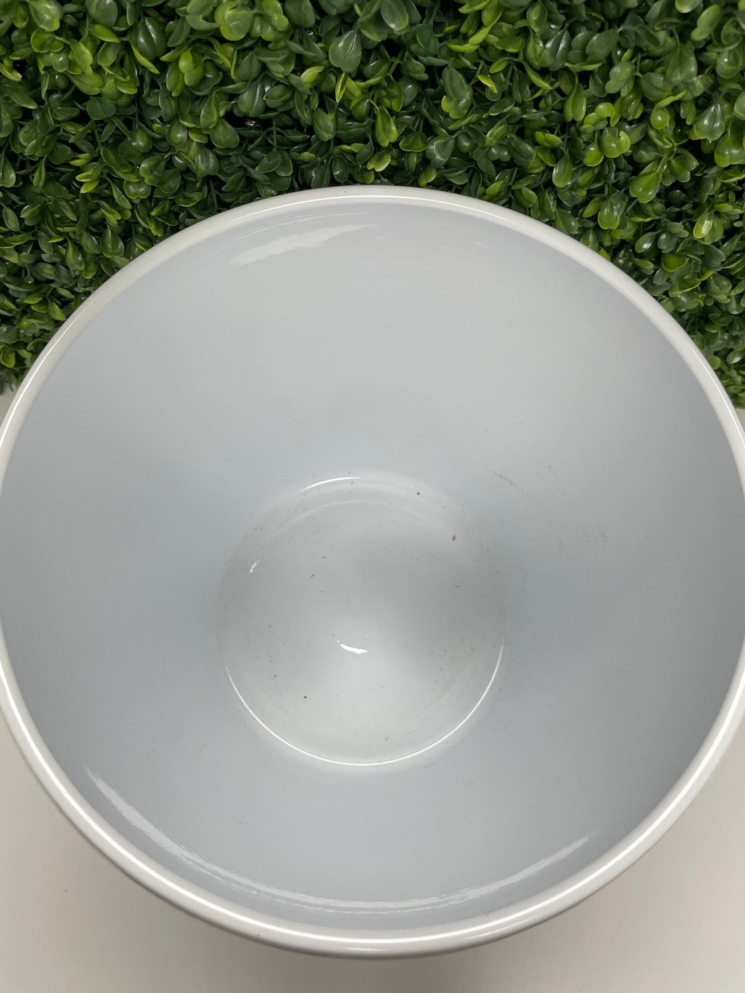 Top-down view of a glossy white ceramic planter, showcasing its smooth interior. The background features green artificial foliage, enhancing the natural aesthetic.