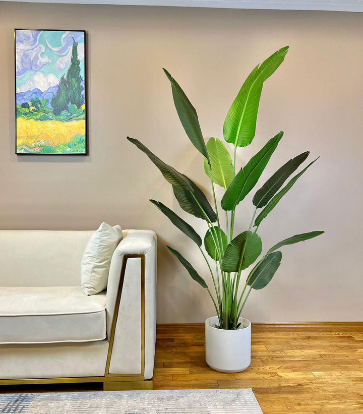 A tall artificial bird of paradise plant in a sleek white planter is placed on a hardwood floor next to a modern cream-colored sofa with gold accents. The plant's large, vibrant green leaves create a striking contrast against the neutral beige wall. A colorful framed painting depicting a scenic landscape adds a touch of artful elegance to the space.