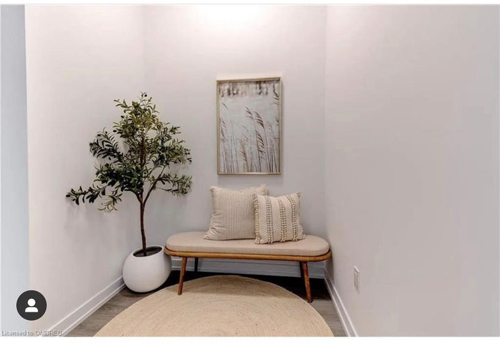A small nook decorated with a minimalist aesthetic. An artificial olive tree in a white round pot is placed on the left, next to a simple wooden bench with two beige cushions. Above the bench hangs a framed art print depicting reeds in soft, neutral tones. The space is complemented by a round, woven rug on the floor, creating a cozy and inviting atmosphere.