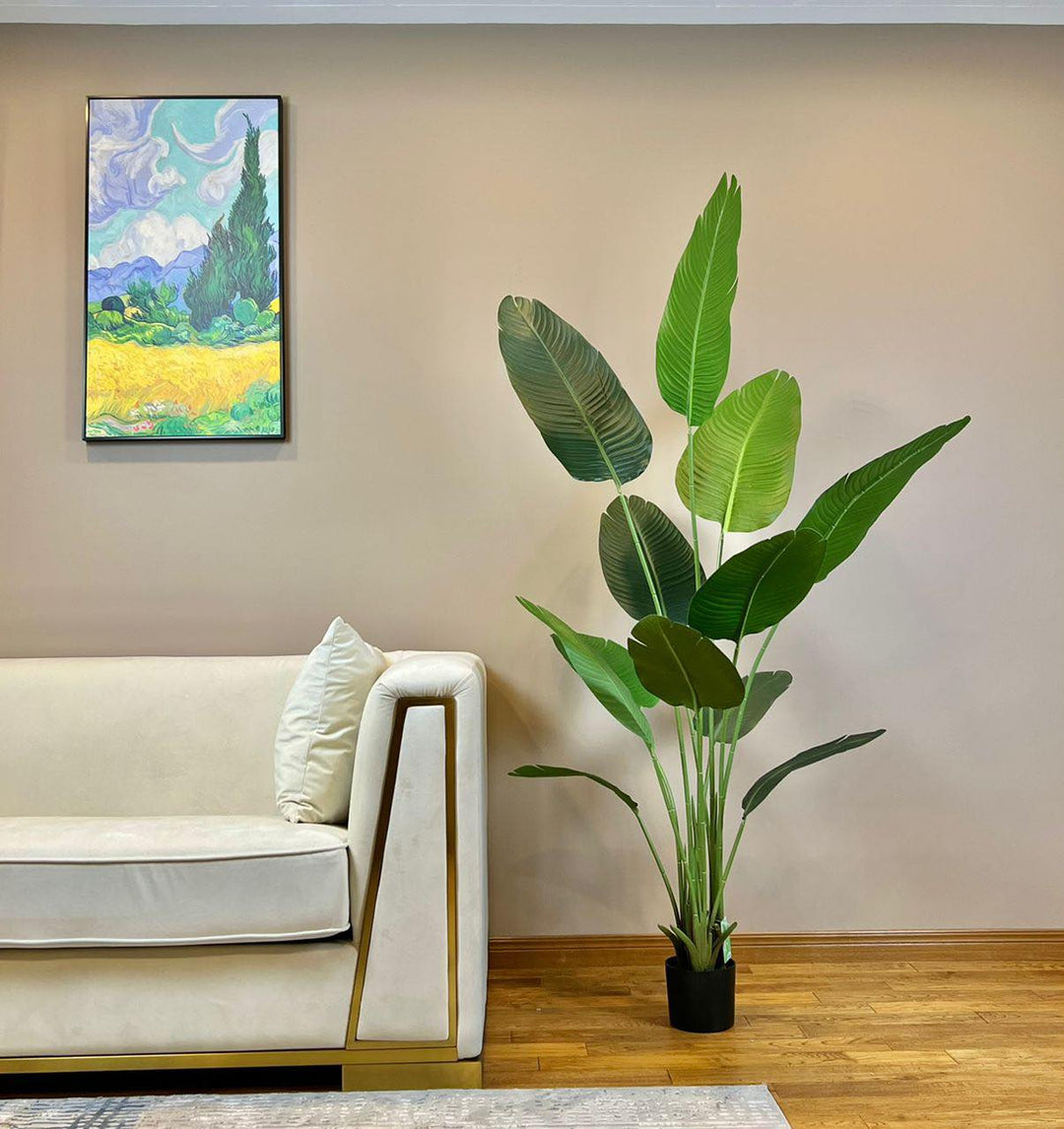 Artificial bird of paradise plant with large green leaves in a black pot placed next to a beige sofa with gold accents in a contemporary living room setting