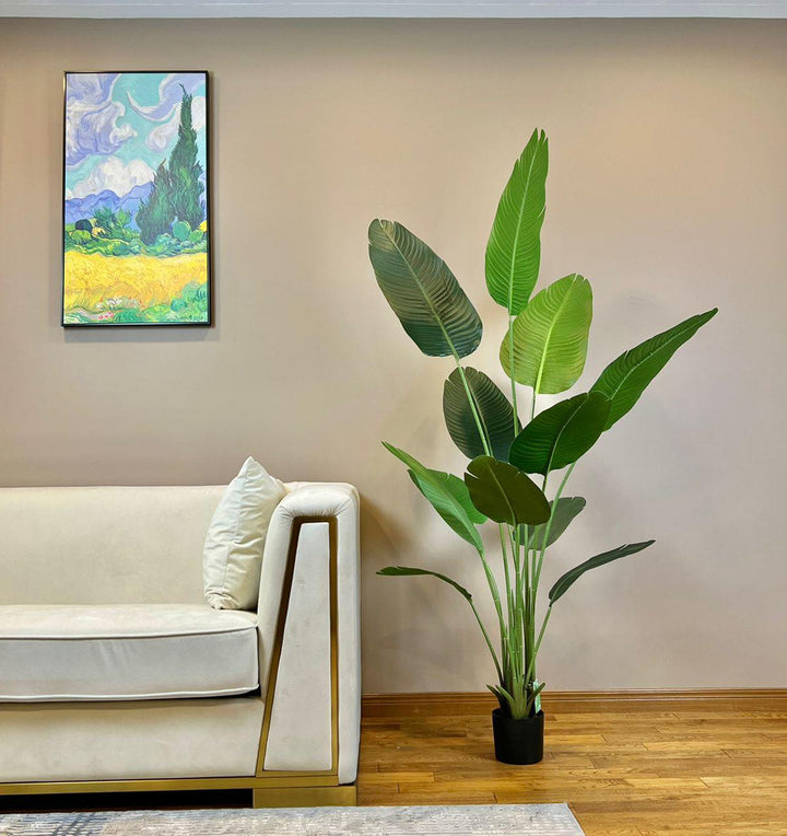 Artificial bird of paradise plant with large green leaves in a black pot placed next to a beige sofa with gold accents in a contemporary living room setting