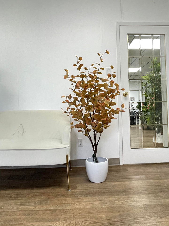 A contemporary indoor space featuring a minimalist setup with a white armchair with gold accents next to a white ceramic planter holding an artificial tree with autumn-colored leaves. The setting includes a wooden floor and a glass door reflecting greenery from another room.
