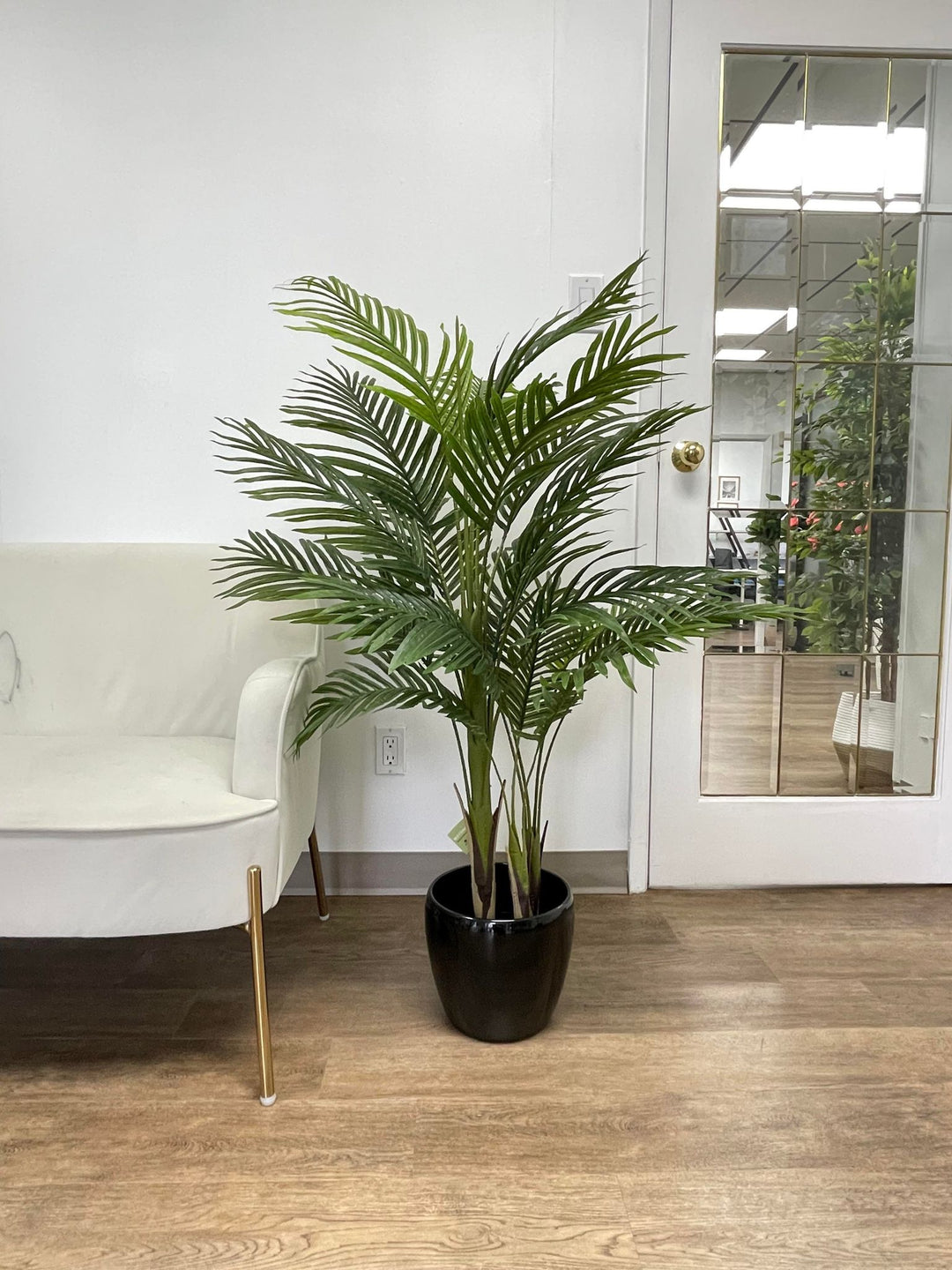 Artificial palm tree in a black ceramic planter next to a white chair, placed indoors with a wooden floor and reflective door in the background.
