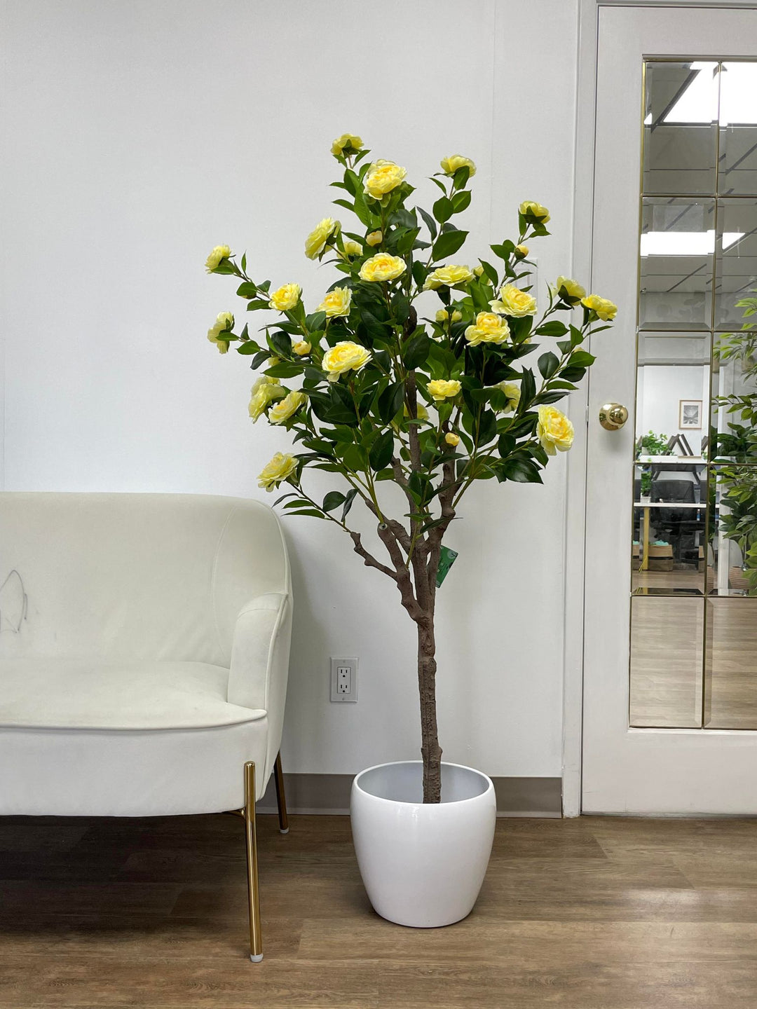 Artificial yellow flower tree in a sleek white pot, positioned beside a modern white chair in a bright and minimalistic room, enhancing the space with a touch of natural beauty.
