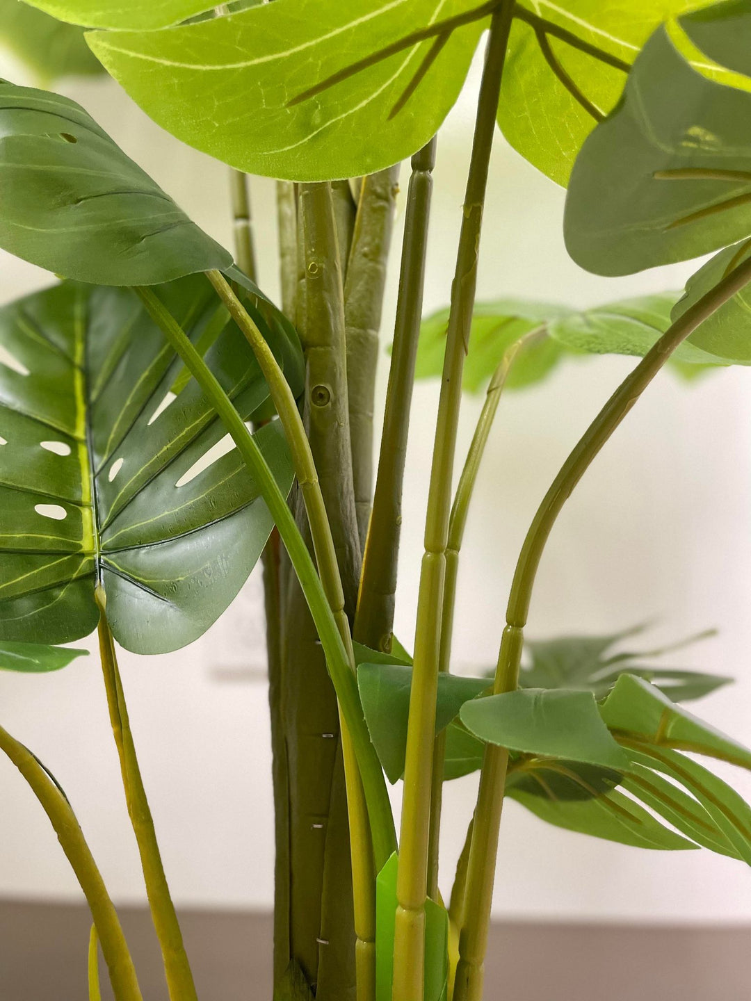 Close-up view of artificial monstera plant stems, featuring vibrant green shades and a natural texture. The image captures the details of the stems with a blurred foreground leaf, adding depth and realism to the plant's appearance
