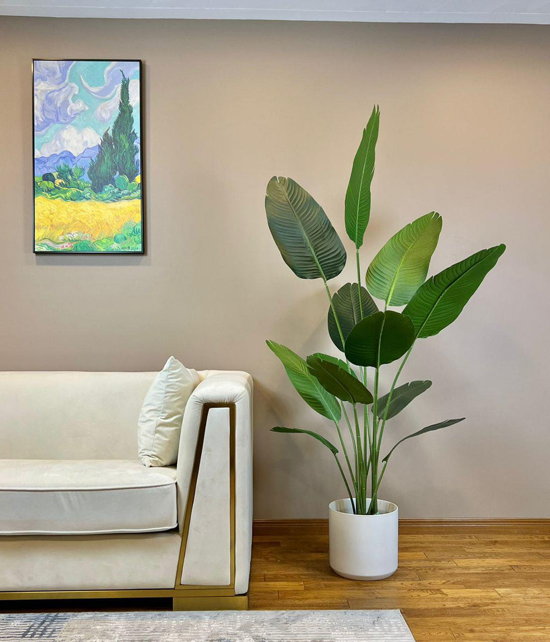 Modern living room with a tall artificial bird of paradise plant in a white pot next to a cream sofa with gold accents and a colorful landscape painting on the wall
