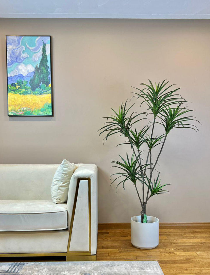 Artificial Dracaena plant in a modern white planter placed beside a luxurious white sofa with gold accents, enhancing the contemporary decor of a living room with wooden flooring and wall art.
