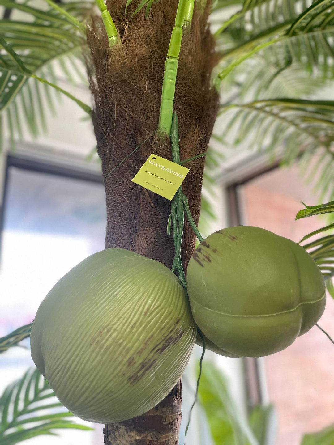 The image shows a close-up of the trunk of an artificial palm tree with two attached coconuts. The coconuts are made from a material that mimics the texture and color of real green coconuts. Attached to the trunk is a small tag labeled "WAYSAVING," likely indicating the brand or manufacturer of the artificial palm tree. The background is slightly blurred, showing what appears to be a bright, indoor setting with large windows.